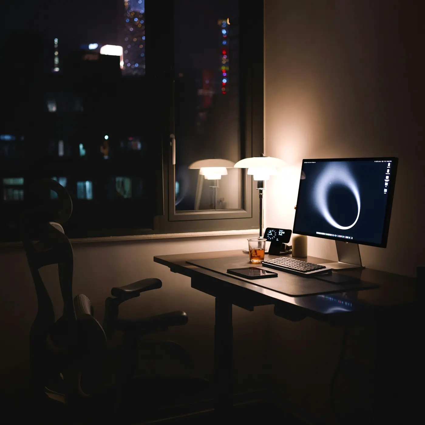 Mesa de trabalho à noite com monitor e luminária acesa, diante da janela com as luzes da cidade