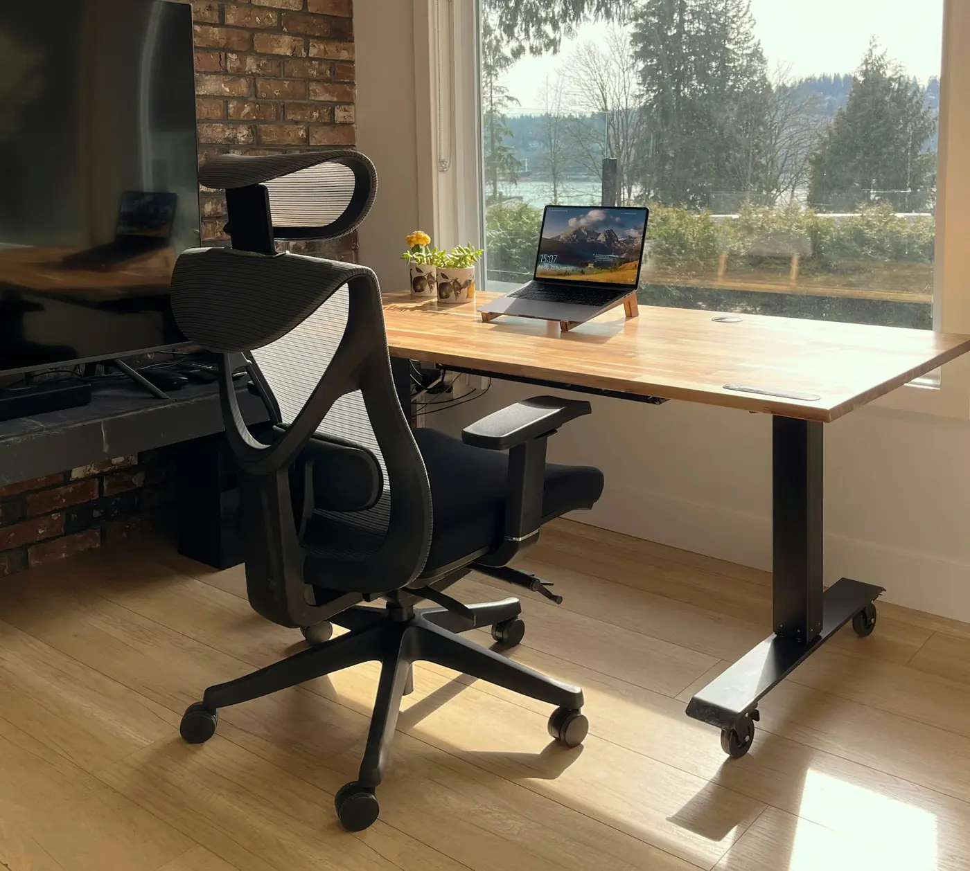 Mesa de trabalho com notebook diante de um janelão, ao lado de uma parede de tijolos aparentes